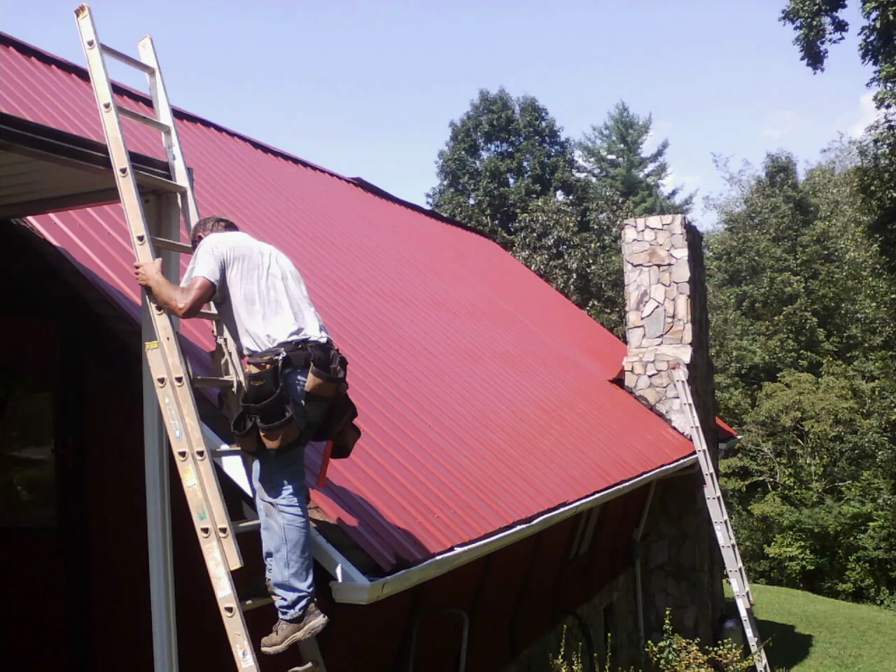 Roofer on a ladder beside a red metal roof and stone chimney — Soffit & Fascia Repair by Roofing Brothers in Maywood